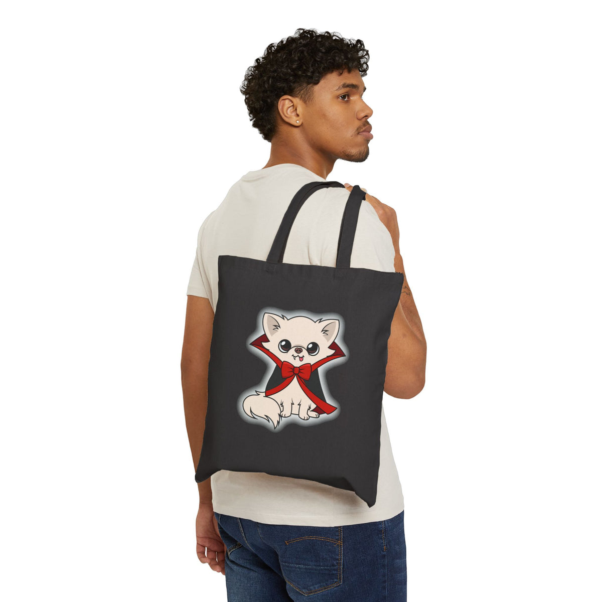 Black cotton tote bag shown over a person’s shoulder, featuring a cream chihuahua sitting calmly in a black cape with red lining and a red bow, outlined by a soft white glow on the front.