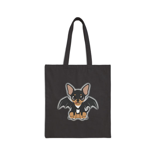 Black canvas tote bag with a centered cartoon of Maya the black-and-tan chihuahua, shown sitting facing forward with a small white chest patch, one tiny fang showing, and dark bat wings outlined in a soft white glow.
