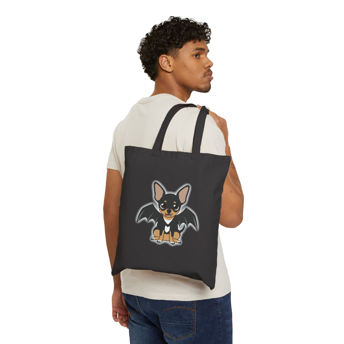 Black canvas tote bag over a person’s shoulder, featuring a cute black-and-tan chihuahua with bat wings, a small fang, and a soft gray glow around the character on the front.