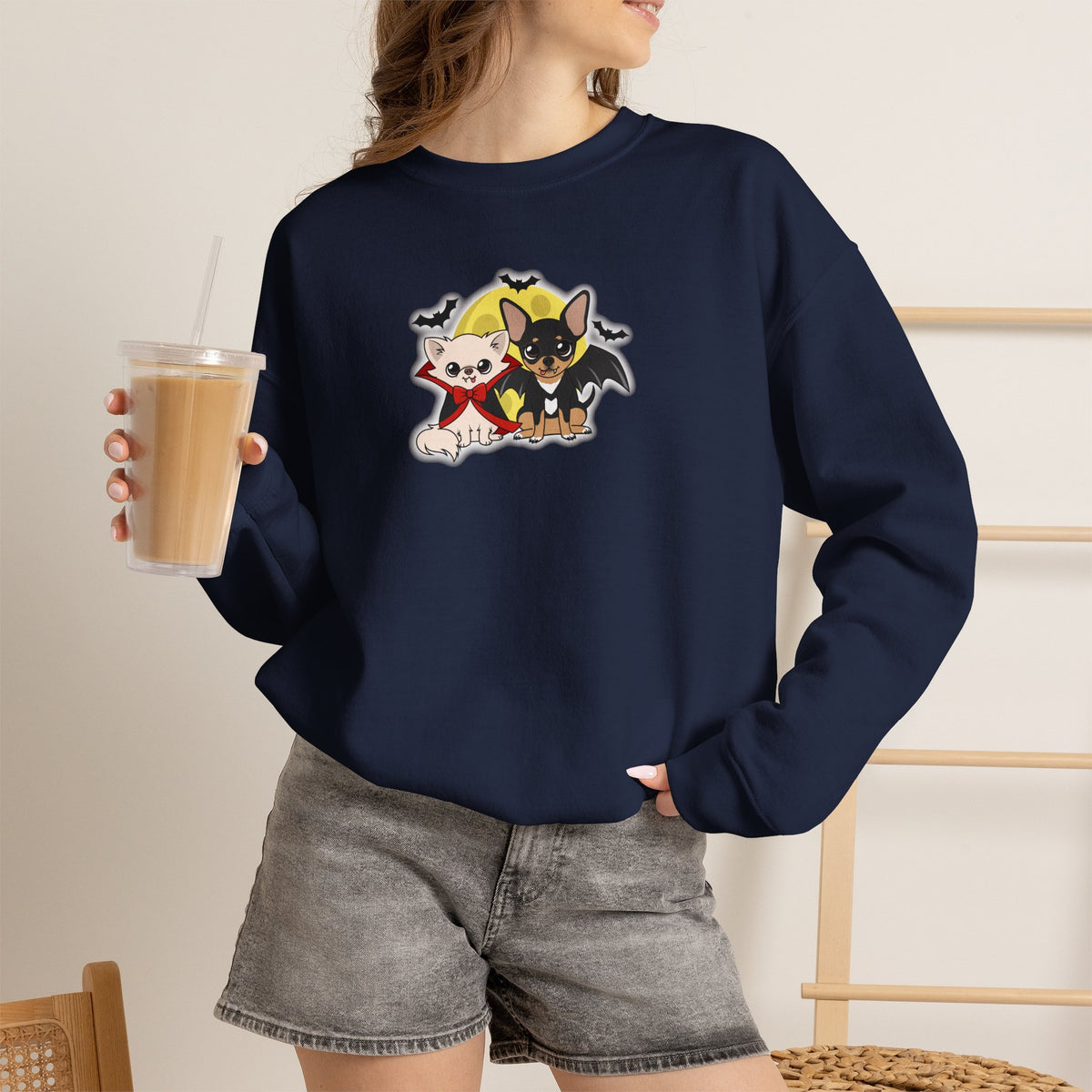 Person wearing a navy crewneck sweatshirt with a centered illustration of two cartoon chihuahuas in gentle vampire costumes, standing side by side in front of a yellow moon with small bats flying above.
