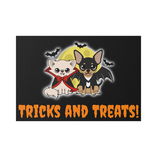 Rectangular black lawn sign with a cartoon cream chihuahua in a red vampire cape and bow beside a black-and-tan chihuahua with bat wings, sitting in front of a yellow moon with small bats flying around, above the orange text that reads “TRICKS AND TREATS!”.