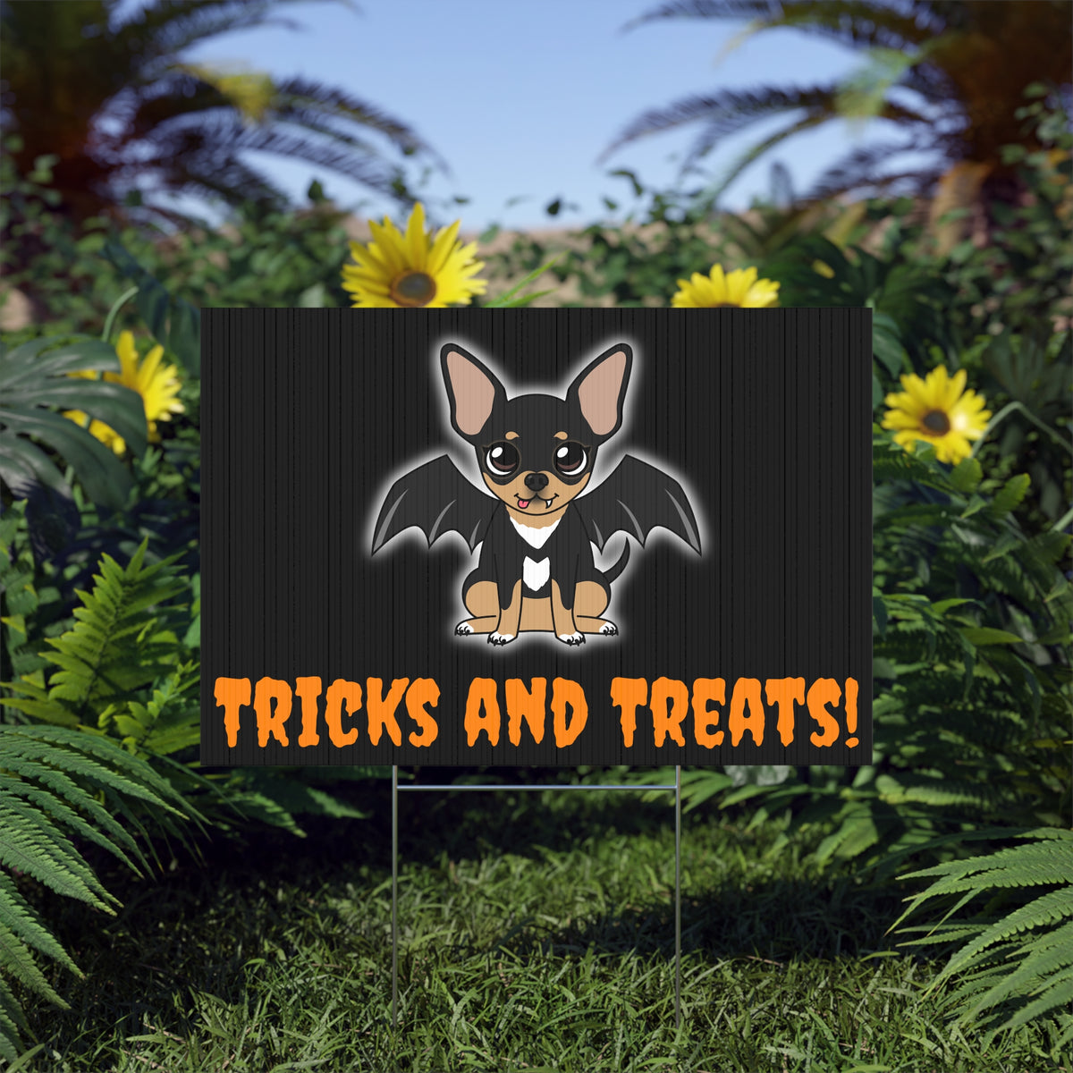 Halloween lawn sign in a garden, showing a cartoon black-and-tan chihuahua with bat wings and a white chest glow on a dark panel background, with bold orange text reading “TRICKS AND TREATS!” along the bottom.