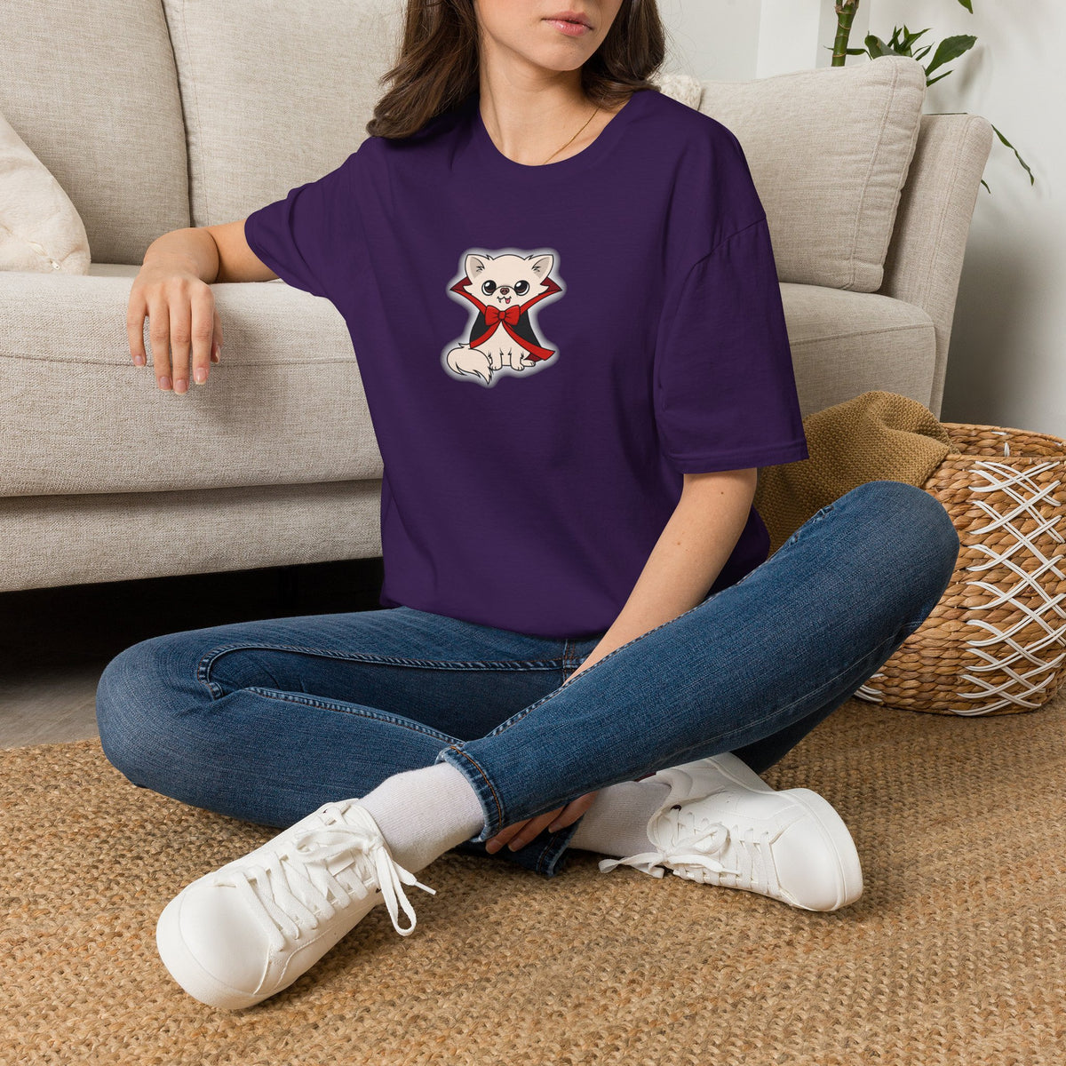 Woman sitting cross-legged on a living room floor wearing a purple t-shirt with a cute cream chihuahua character in a black cape with red lining and a red bow printed on the front.