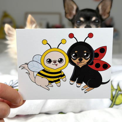 A hand holds a square white card featuring a cute cartoon illustration of two small dogs dressed as a bee and ladybug, with a real dog blurred in the background in a bedroom setting.