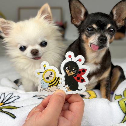Two small dogs sitting on a white blanket with floral embroidery, while a human hand holds up two cartoon stickers of a bee and a ladybug in front of them.