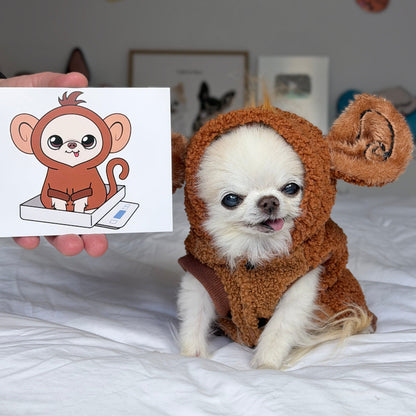 A small white dog wearing a brown plush monkey costume sits on a white bed, while a hand holds up a printed card showing a cartoon illustration of a monkey sitting on a box.