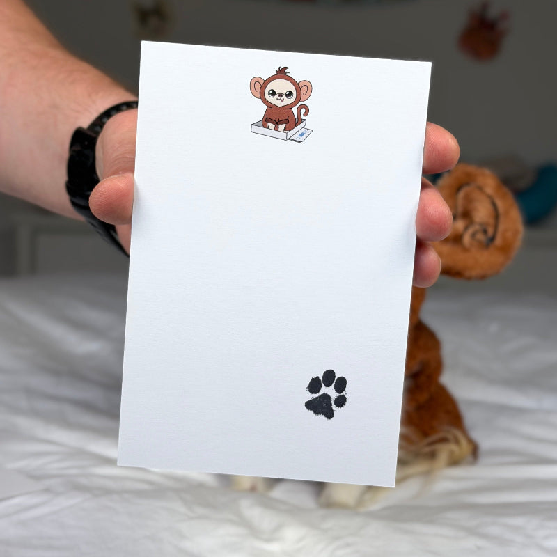 A hand holds a white rectangular card or envelope in front of a bedroom background. The card has a small cartoon monkey illustration near the top and a black paw print near the bottom.
