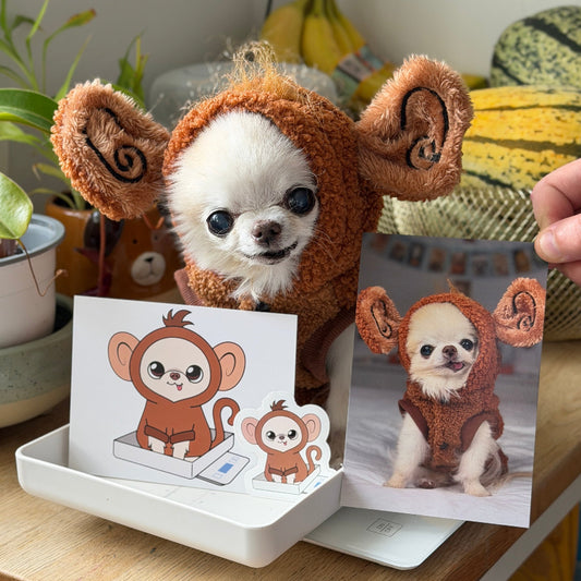 A small white and tan dog wearing a brown plush animal costume sits on a wooden table, with a printed card and photo of the dog in costume displayed in front. A human hand is visible holding the printed photo on the right.
