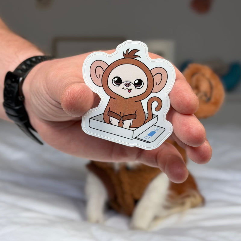 A hand holds a die-cut sticker featuring a cute cartoon brown monkey sitting on a white scale, with a blurred cat and bed in the background.