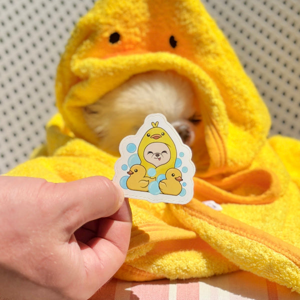 A small fluffy white dog wrapped in a bright yellow duck-shaped hooded towel sits on a white slatted outdoor chair, while a human hand holds a cute cartoon sticker in front of the dog's face.