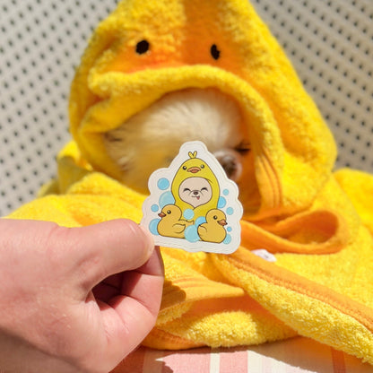 A small fluffy white dog wrapped in a bright yellow duck-shaped hooded towel sits on a white slatted outdoor chair, while a human hand holds a cute cartoon sticker in front of the dog's face.