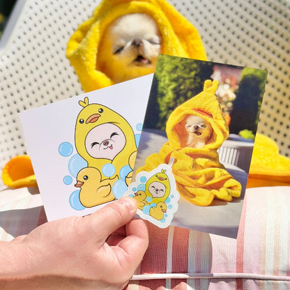 A hand holds two printed stickers featuring a cute cartoon duck in a yellow hooded outfit, with a real dog wearing a matching yellow hooded towel sitting in the background on a white outdoor chair.