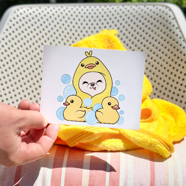 A hand holds a small printed card featuring a cute cartoon duck character in a yellow hood with blue bubbles, set against a yellow towel and striped cushion in an outdoor patio setting.