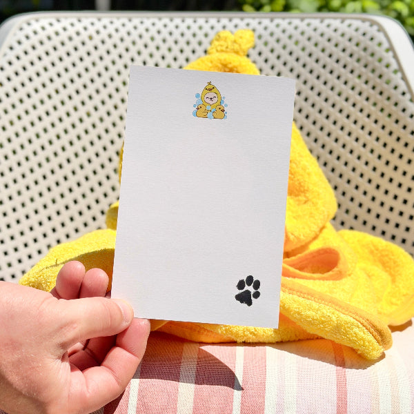 A hand holds a white rectangular card featuring a small cartoon character near the top and a black paw print near the bottom, set against a yellow towel and pink-and-white striped cushion in an outdoor chair.