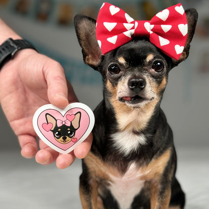 A hand holding Maya's Happy Heart Sticker next to Tiny Chihuahua Maya, who is wearing a heart bow on her head, highlighting the sticker's design with pink hearts alongside the real-life muse, creating a charming and affectionate scene.