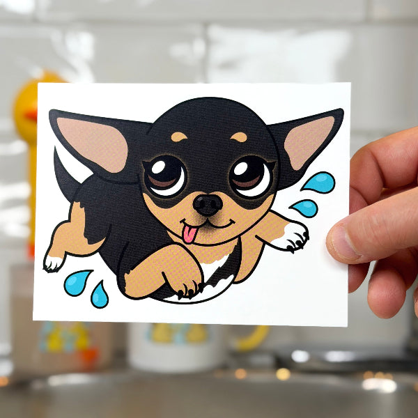 A hand holds a square white sticker or card featuring a cartoon chihuahua puppy with a black and tan winter hood, set against a blurred bathroom sink background.