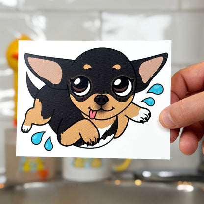 A hand holds a square white sticker or card featuring a cartoon chihuahua puppy with a black and tan winter hood, set against a blurred bathroom sink background.