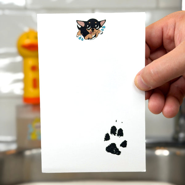 A hand holds a white rectangular card in a kitchen setting, featuring a small cartoon dog illustration near the top and a black paw print near the bottom.