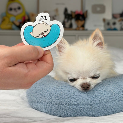 A hand holds a die-cut sticker featuring a cartoon dog sleeping in a blue pillow with 'Z' marks above its head, next to a small fluffy white dog resting on a blue cushion in a bedroom setting.