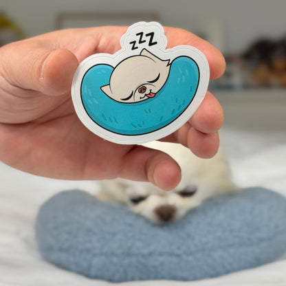 A hand holds a die-cut sticker featuring a cartoon cat sleeping in a blue crescent pool with 'Z Z Z' above its head. In the blurred background, a small white dog rests on a blue cushion on a bed.