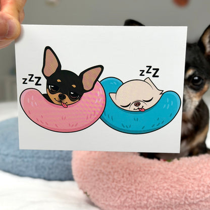 A hand holds a white card featuring a cartoon illustration of two sleeping dogs in pink and blue crescent-shaped beds with 'zzz' text. A real dog is visible in the background on a bed with pink and blue pillows.