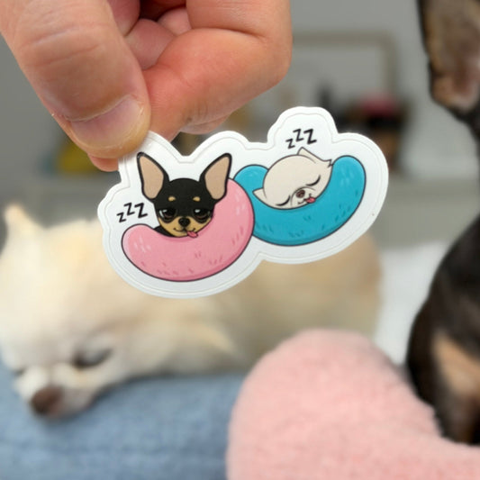 A hand holds a die-cut sticker featuring two cartoon dogs sleeping in pink and blue necklaces, with blurred dogs and a pet bed in the background.