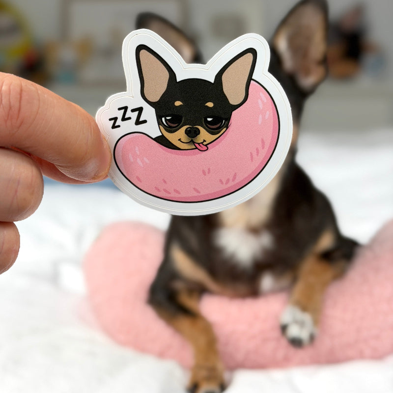 A hand holds a die-cut sticker featuring a cartoon chihuahua sleeping in a pink donut, with 'zzz' marks, while a real chihuahua sits in the blurred background on a bed.