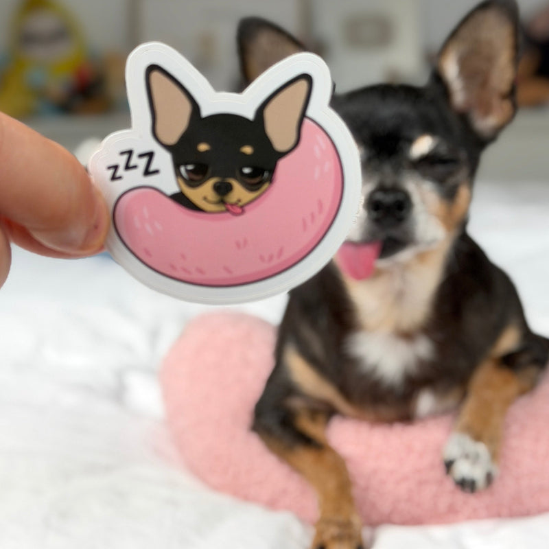 A hand holds a die-cut sticker featuring a cartoon Chihuahua sleeping in a pink donut, with 'Z Z Z' marks, while a real Chihuahua dog sits in the background on a pink cushion.