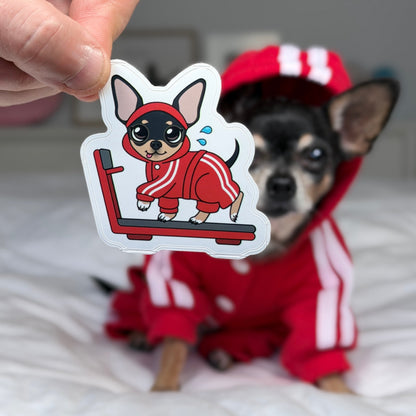 A hand holds a die-cut sticker featuring a cartoon chihuahua running on a treadmill in a red tracksuit, with a real chihuahua wearing a matching red tracksuit in the blurred background on a white bed.
