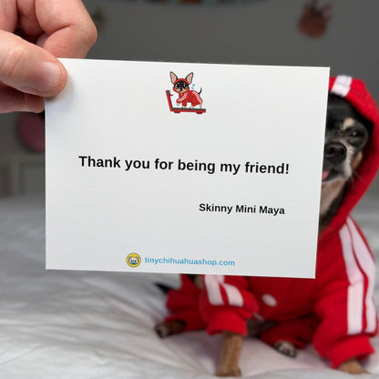 A hand holds a white printed card that reads 'Thank you for being my friend! Skinny Mini Maya' with a small chihuahua illustration. A real dog wearing a red outfit sits on a white bed in the background, creating a warm, personalized scene.