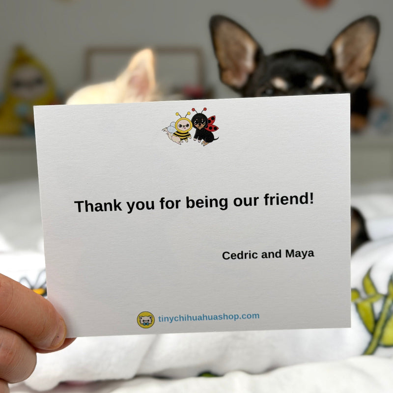 A hand holds a white personalized thank you card in front of a black dog and a white cat in a cozy bedroom setting. The card features a cute cartoon bee and ant design at the top and the text 'Thank you for being our friend! Cedric and Maya' with the website 'linychihuahuaashop.