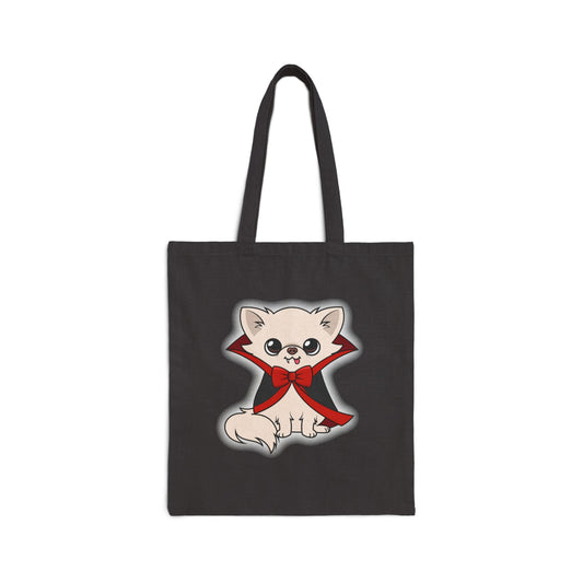 Black canvas tote bag featuring a cute cream-colored chihuahua sitting upright in a black cape with red lining and a large red bow, tiny fangs showing and tongue out, centered on the front with a soft white glow around the character.