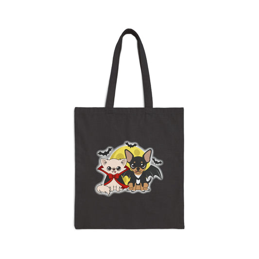 Black cotton tote bag featuring two cartoon chihuahuas in Halloween outfits, one cream dog in a red-lined cape and bow and one black-and-tan dog with bat wings, sitting together in front of a yellow moon with small bats flying around.