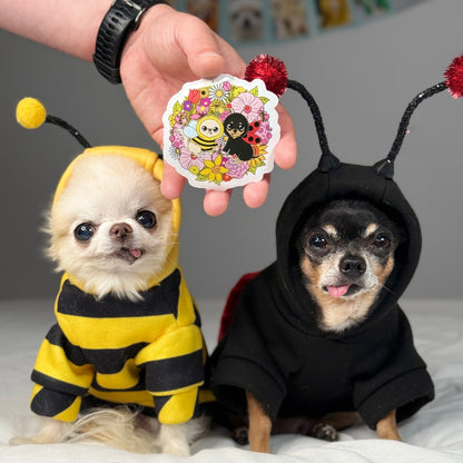 Two small dogs in bee and ladybug black hooded costumes with a person holding a colorful sticker.
