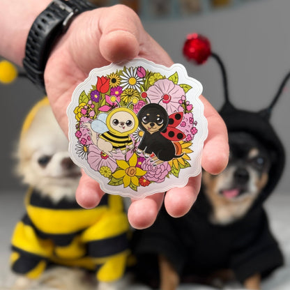 Hand holding an illustrated sticker featuring Cedric as a bee and Maya as a ladybug amid a colorful bouquet of blooming flowers.
