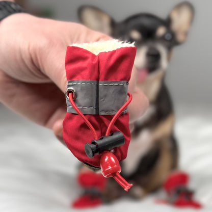 Closeup of the adjustable strap and faux-fur interior of a red small dog boot, held in front of a Chihuahua wearing matching boots.