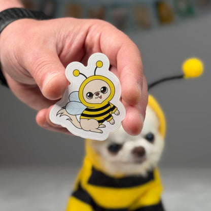 Hand holding an illustrated sticker of Cedric the Chihuahua dressed as a bee with yellow-and-black stripes, wings and antennae.
