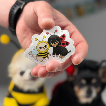 Hand holding an illustrated sticker showing two Chihuahuas; one in a bee costume and the other in a ladybug costume, both with wings.
