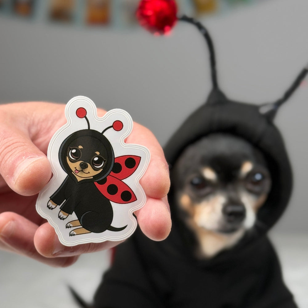 Hand holding an illustrated sticker of Maya the Chihuahua dressed as a flying ladybug with red-and-black wings and antennae.