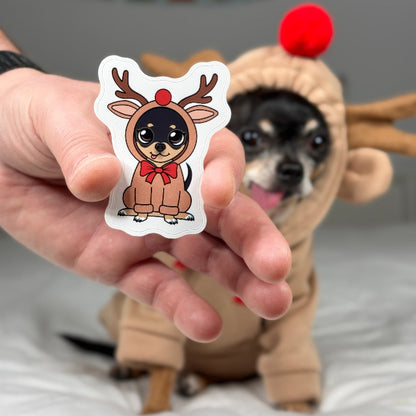 Person holding a sticker of a reindeer dog with a dog in a reindeer costume in the background.