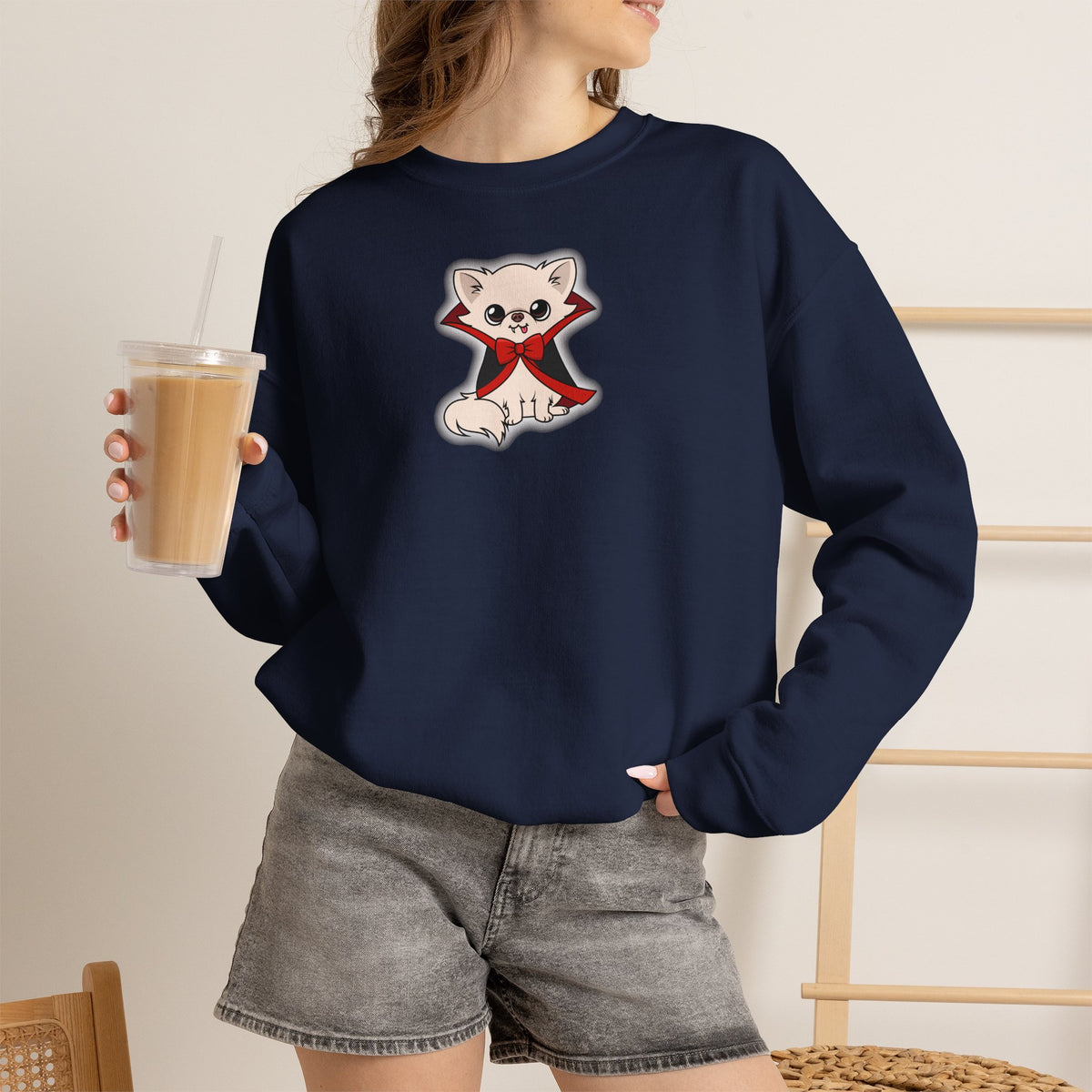 Person wearing a navy crewneck sweatshirt with a centered graphic of a cream chihuahua sitting in a black cape with red lining and a red bow, shown against a soft white glow, holding an iced coffee cup in one hand.